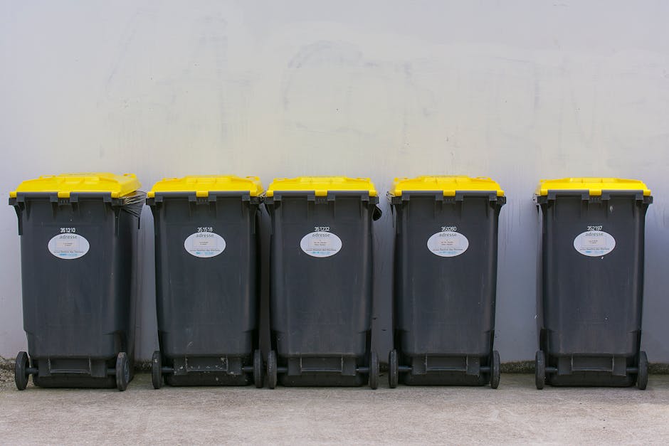 A person's arm and hand are visible, holding a green plastic rubbish bag by its gathered top, which is twisted and tied. The bag appears to contain light-colored waste, and has a shiny surface with so